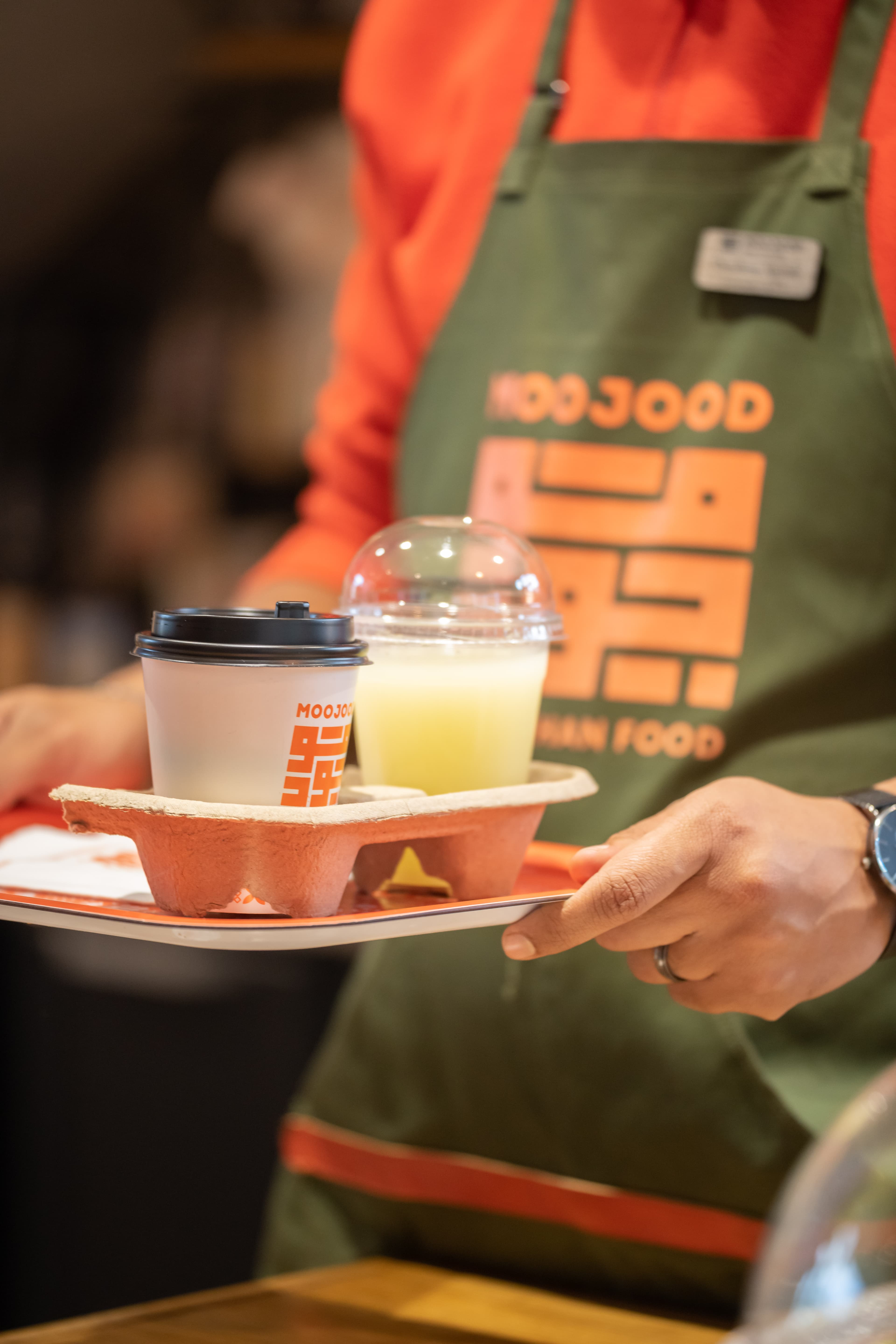 Plateau avec gobelets Moojood : café et jus, porté par un serveur en tablier vert.